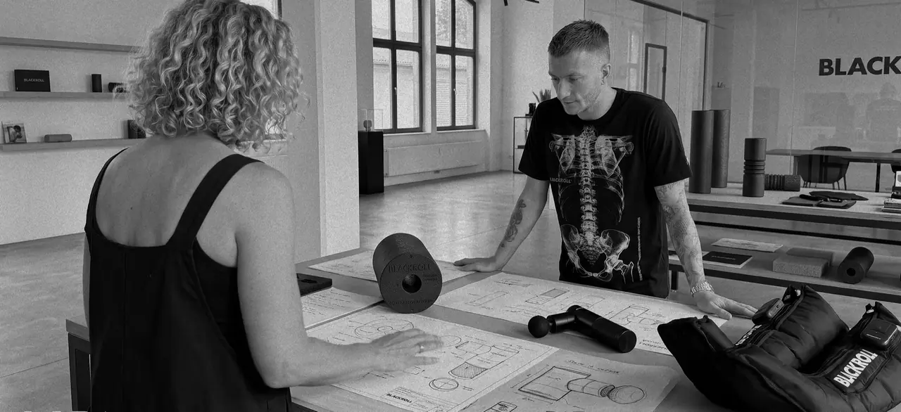Schwarzweißfoto im BLACKROLL LAB: Marco Reus stützt sich auf einen Tisch voller Produktzeichnungen, darauf eine Rolle, eine Fascia Gun und Compression Boots; ihm gegenüber steht eine Kollegin.