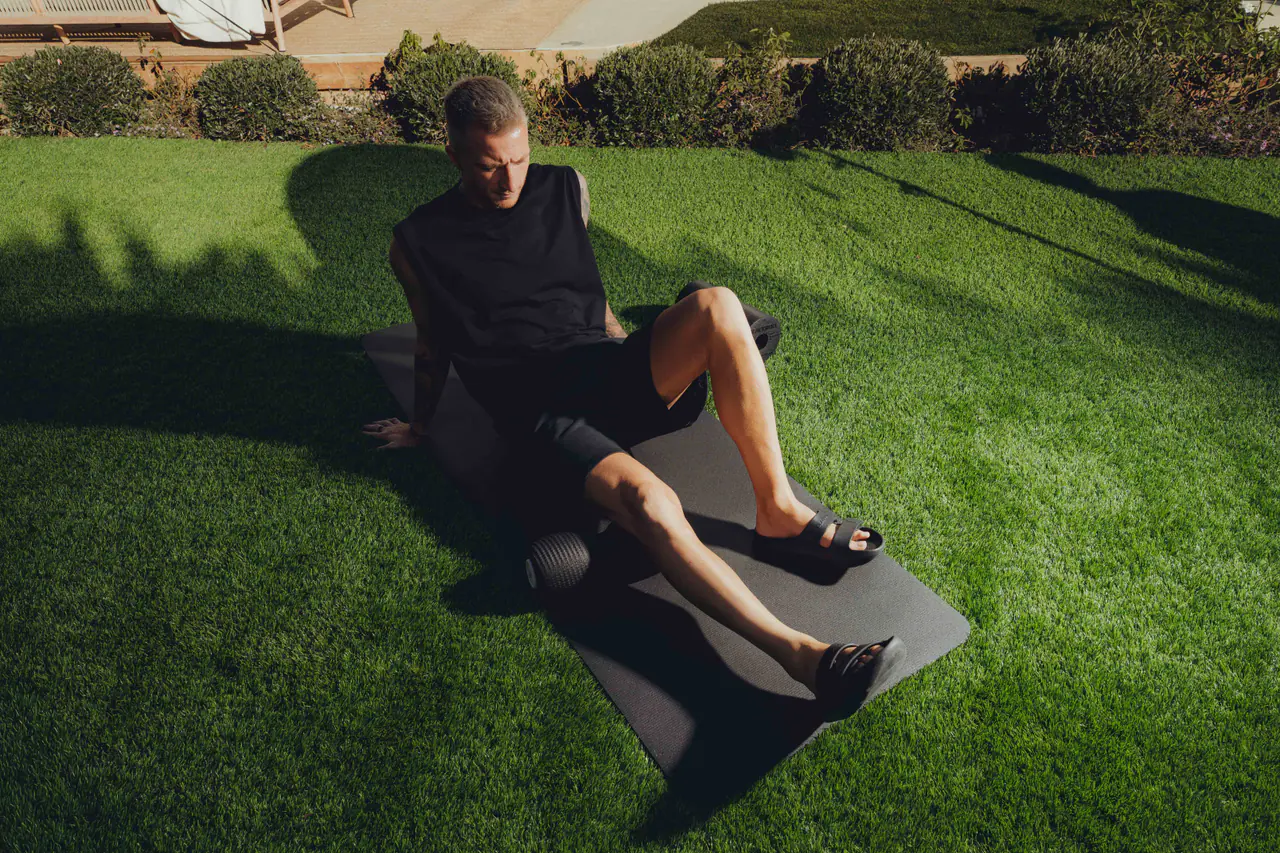 Marco Reus auf einer schwarzen Matte auf dem Rasen bei tief stehender Sonne, ein Bein angewinkelt, Slides an den Füßen, zwei Faszienrollen auf der Matte.