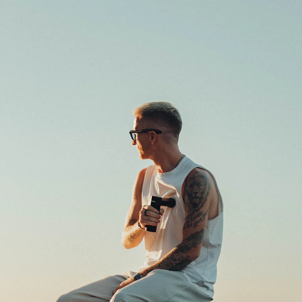 Marco Reus sitzt im Abendlicht vor hellem Himmel, eine Massagepistole in der Hand, und blickt in die Ferne.