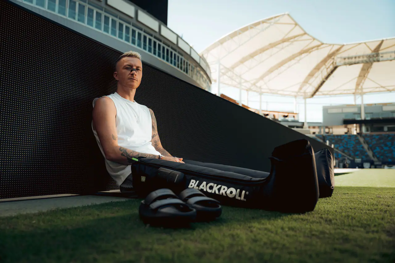 Marco Reus sitzt mit dem Rücken an der LED-Bande auf dem Rasen, beide Beine in BLACKROLL Compression Boots, die Slides vor sich.