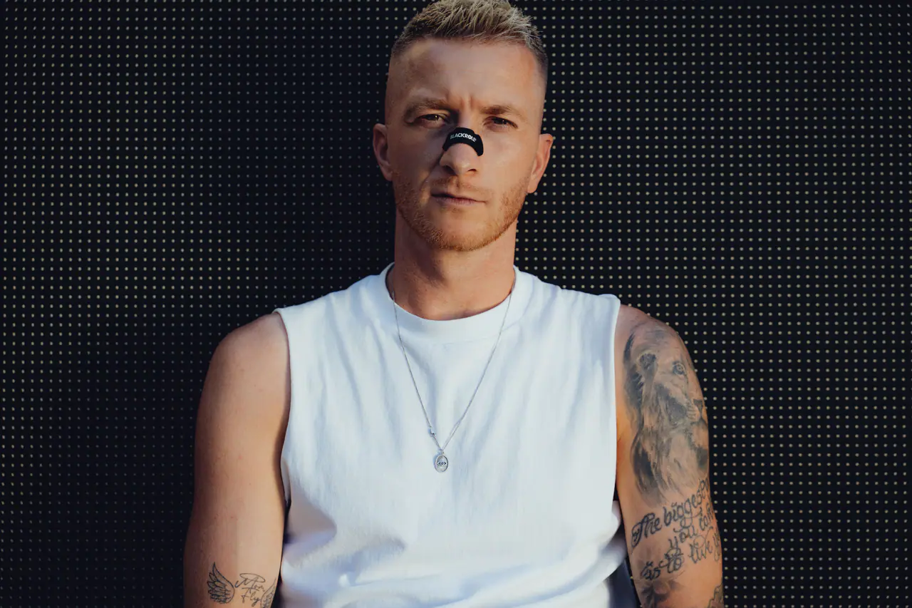 Marco Reus im weißen Tanktop mit Nasenpflaster vor einer schwarzen LED-Wand, der Blick geht direkt in die Kamera.